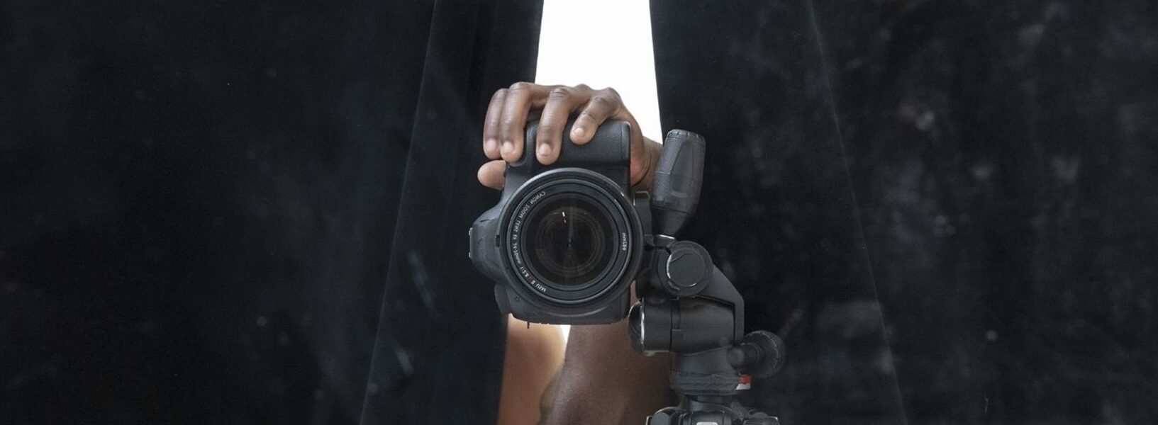 Fotograf:in mit Kamera auf Stativ blickt durch einen Spalt zwischen schwarzen Vorhängen – Blick hinter die Kulissen einer Aufnahme.