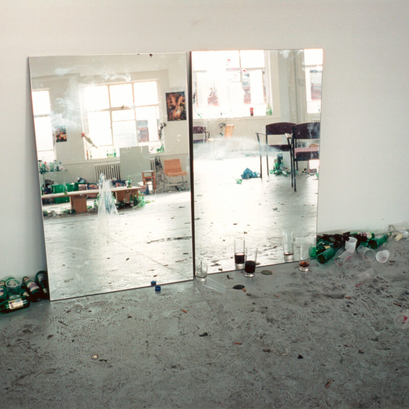 Zwei große Spiegel lehnen an einer weißen Wand, davor liegen viele leere Flaschen und Becher auf dem Boden eines hellen Raums.