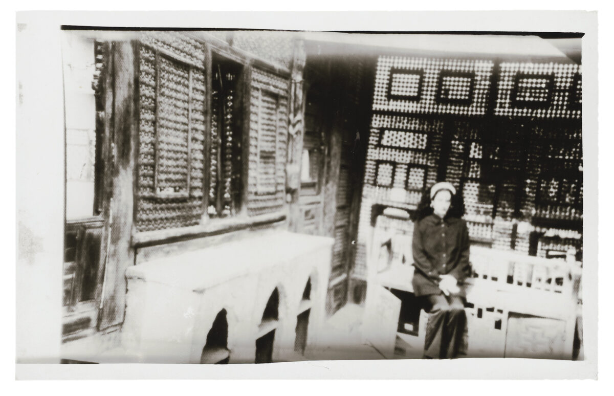 Eine kontrastreiche Schwarz-Weiß-Fotografie im Vintage-Stil, die eine Person zeigt, die in einem Innenhof oder einem Raum mit traditioneller orientalischer Architektur sitzt. Im Hintergrund sind aufwendige Mashrabiya-Holzgitter (gedrechselte Holzfenstergitter) zu sehen. Im Vordergrund befindet sich ein heller Steinsockel mit spitzbogigen Nischen. Die Aufnahme wirkt leicht unscharf und hat den Charakter einer alten Sofortbildaufnahme mit hellem Rand.