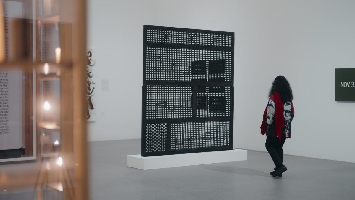 Susan Hefuna mit lockigem Haar und gemusterter Jacke steht in einer Ausstellung vor ihrem Werk 'Knowledge is Sweeter than Honey (Arabic), 2012', einer großformatigen, durchbrochenen Wandstruktur auf Sockel.