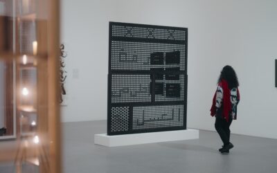 Susan Hefuna mit lockigem Haar und gemusterter Jacke steht in einer Ausstellung vor ihrem Werk 'Knowledge is Sweeter than Honey (Arabic), 2012', einer großformatigen, durchbrochenen Wandstruktur auf Sockel.