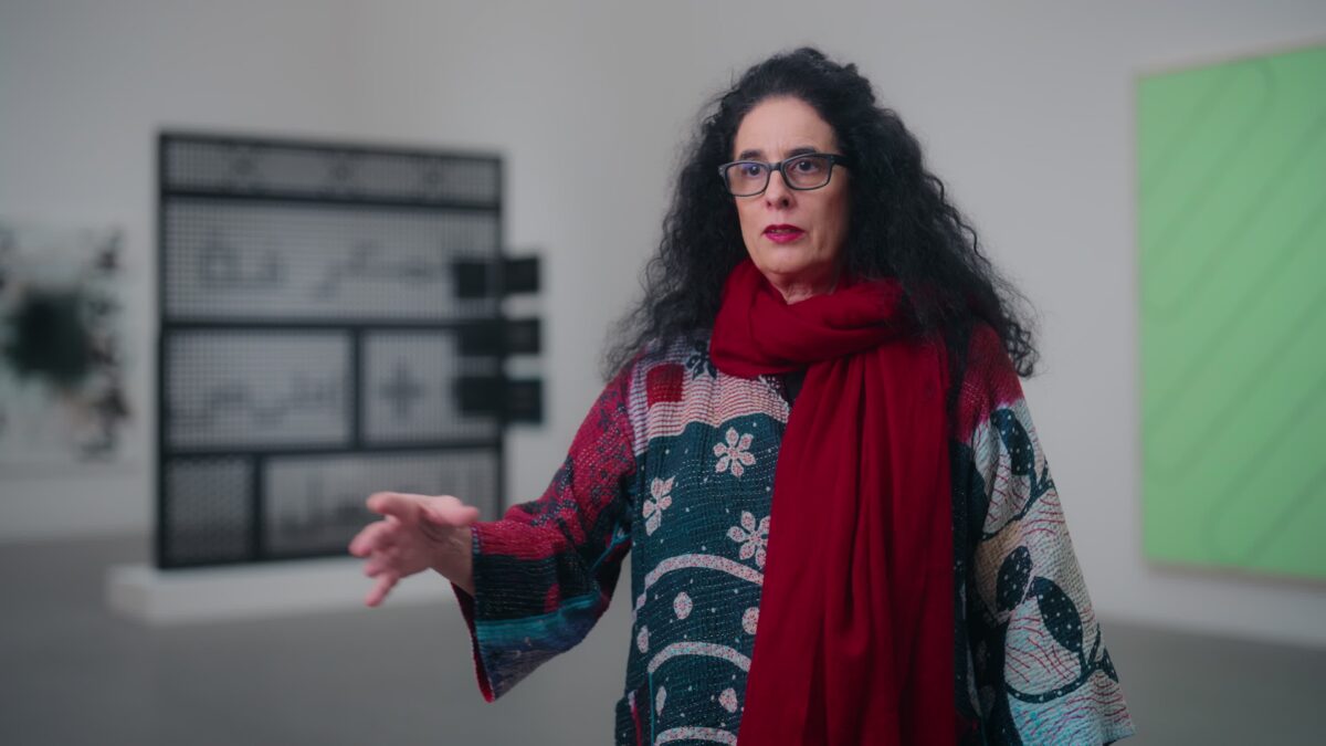 Person mit langen dunklen Haaren trägt einen bunten Pullover mit Mustern und einen roten Schal in einem Raum mit Kunstwerken an den Wänden