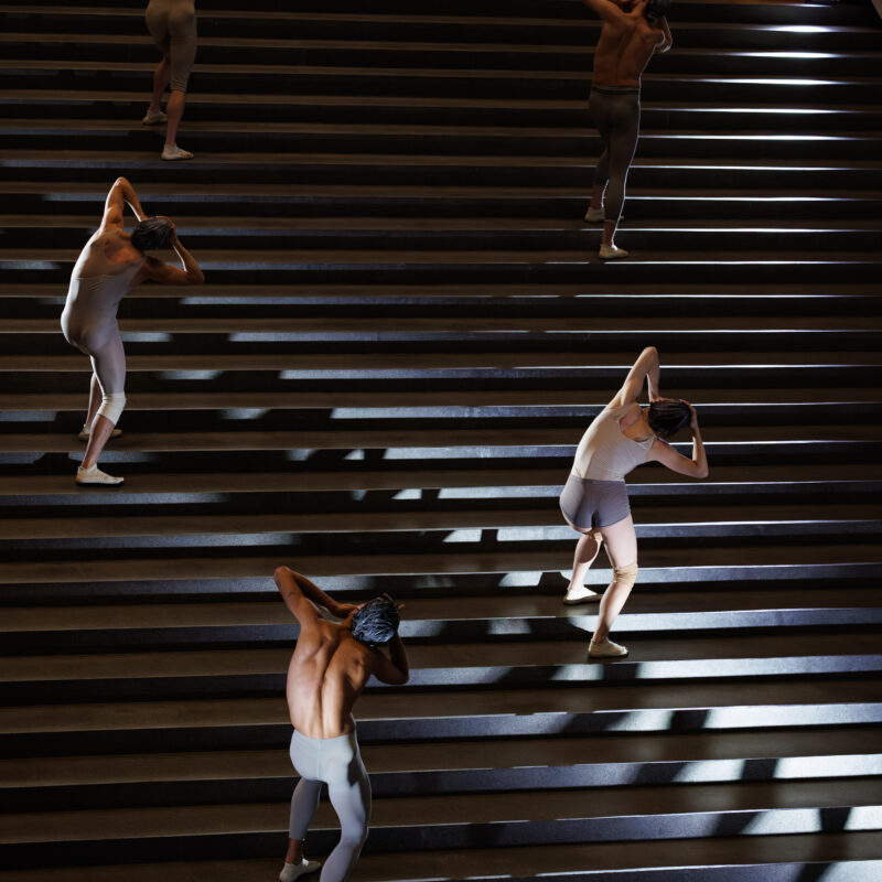 Sechs Tänzer in hautfarbenen Trikots bewegen sich auf einer breiten Treppe, jeweils mit erhobenen Armen hinter dem Kopf.