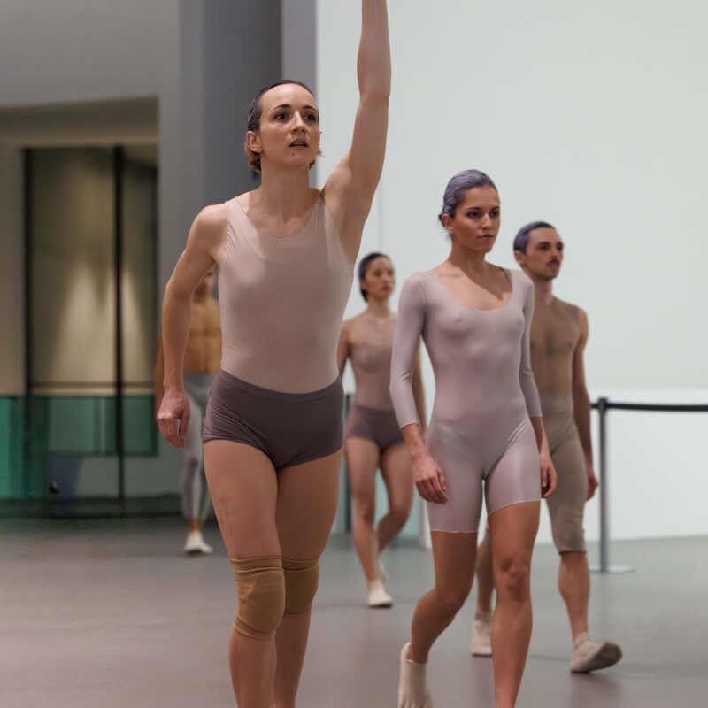 Mehrere Personen in enganliegender Tanzkleidung gehen aufrecht durch einen hellen Raum, eine Person hebt den rechten Arm.