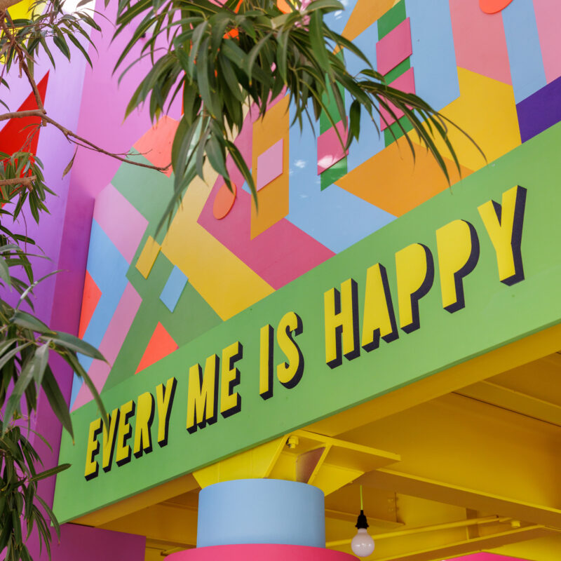 Bunte Wand mit geometrischem Muster und gelbem Schriftzug 'EVERY ME IS HAPPY' über einem pinken Säulenabschnitt und grünen Blättern im Vordergrund