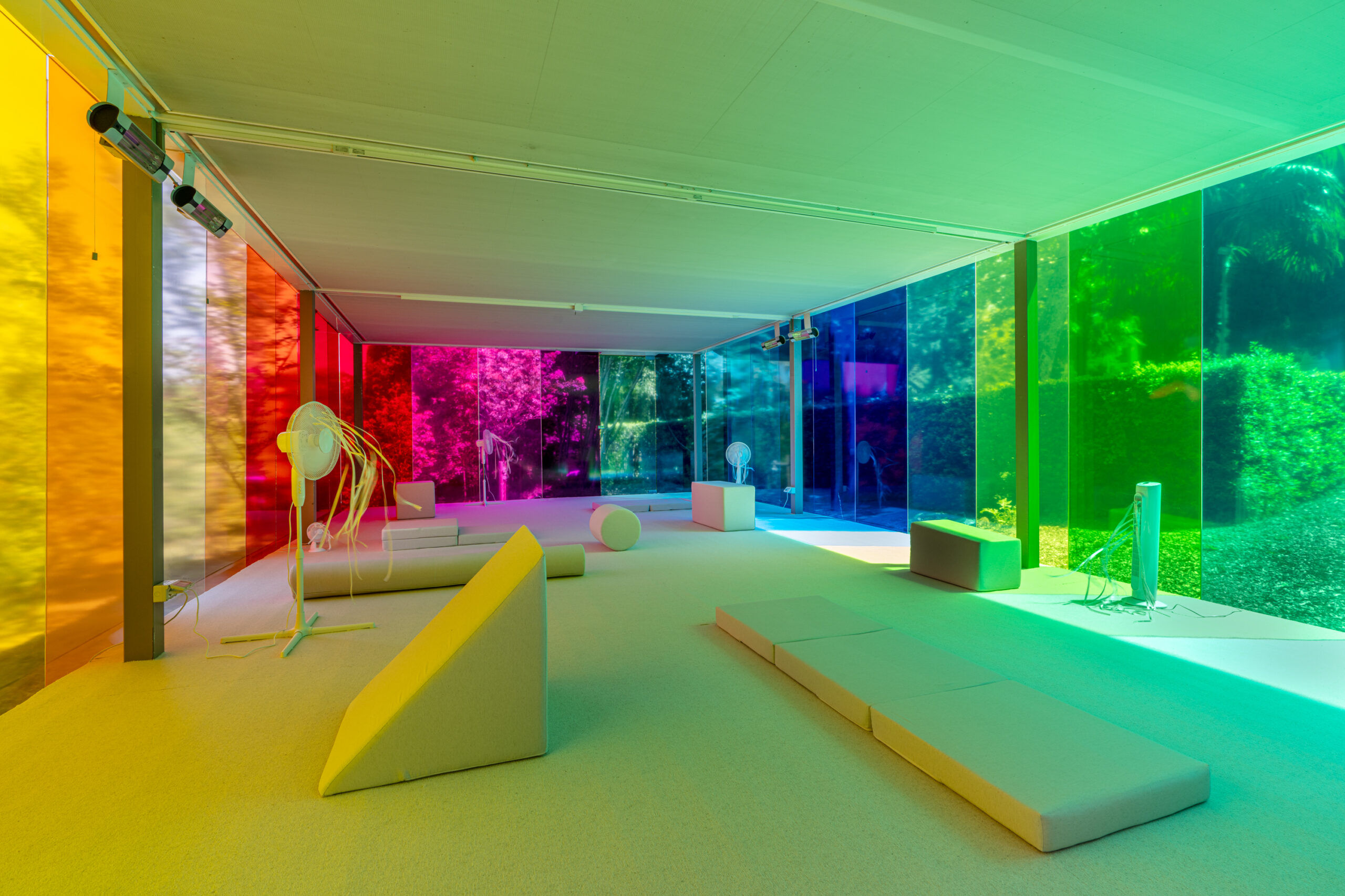 Großer Raum mit geometrischen Sitzmöbeln und Skulpturen, dessen Fenster in Regenbogenfarben getönt sind