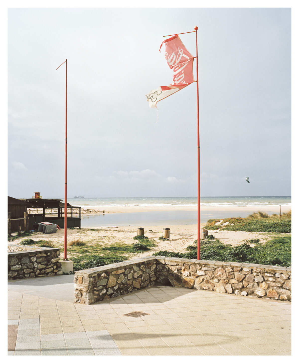 Zwei rote Fahnenmasten mit einer zerrissenen Fahne vor einer Strandlandschaft mit niedrigen Steinmauern und einem Gebäude im Hintergrund.