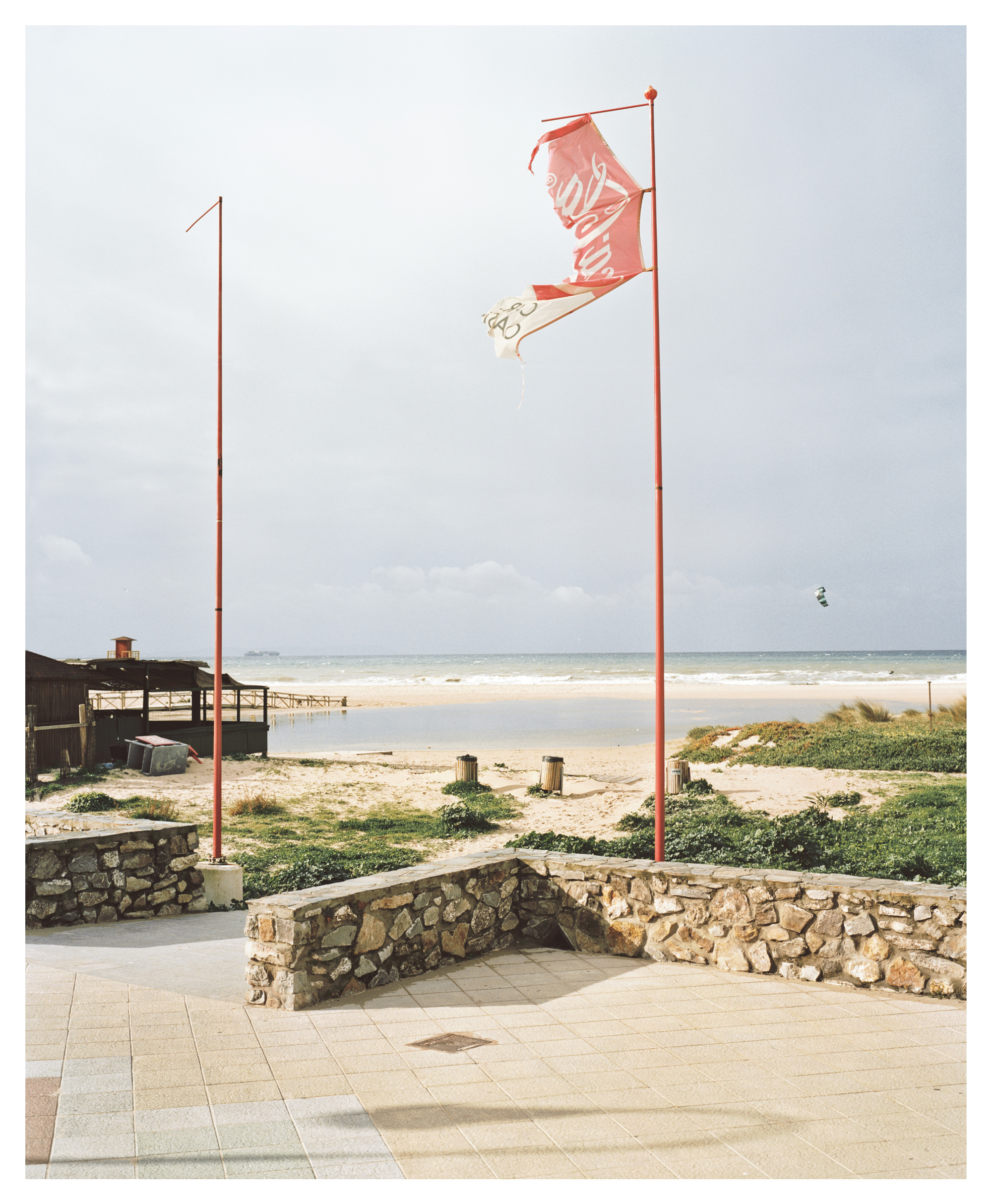 Zwei rote Fahnenmasten mit einer zerrissenen Fahne vor einer Strandlandschaft mit niedrigen Steinmauern und einem Gebäude im Hintergrund.